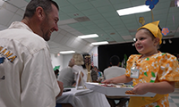  A young girl wearing a floral dress serves a platter of food to a parent at a school. 